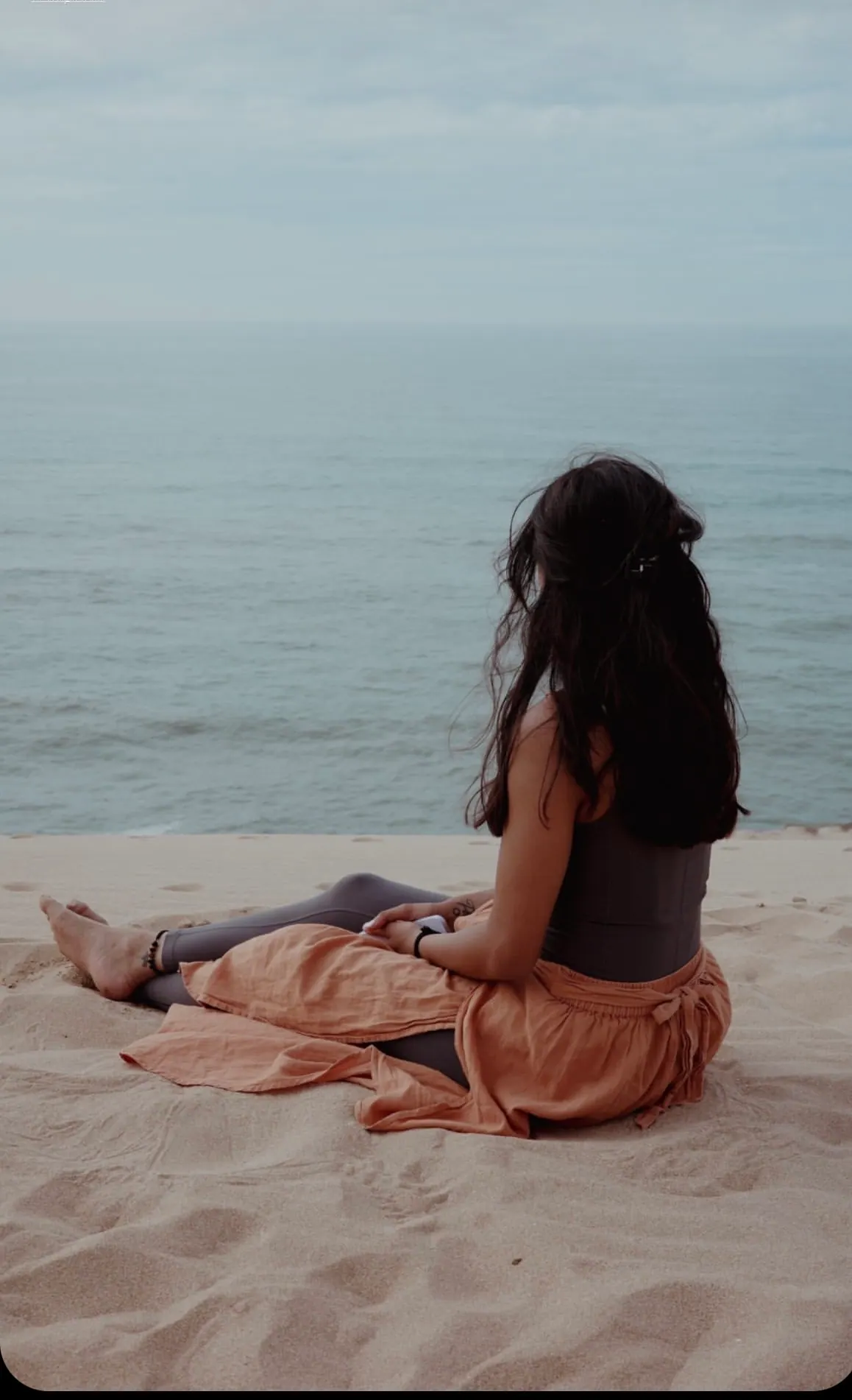 Woman seated on a sandy dune looking out over a calm sea